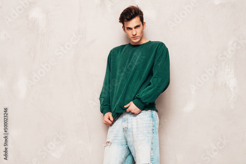 Portrait of handsome confident stylish hipster lambersexual model.Man dressed in green sweater and jeans. Fashion male posing in studio near grey wall