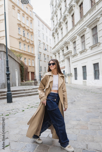 Photography full length of cheerful young woman in trendy sunglasses and trench coat walking on street of vienna