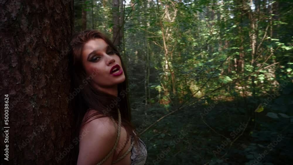 Two women in underwear are tied with a rope to a tree in the woods. Two women in underwear are tied with a rope to a tree in the woods.