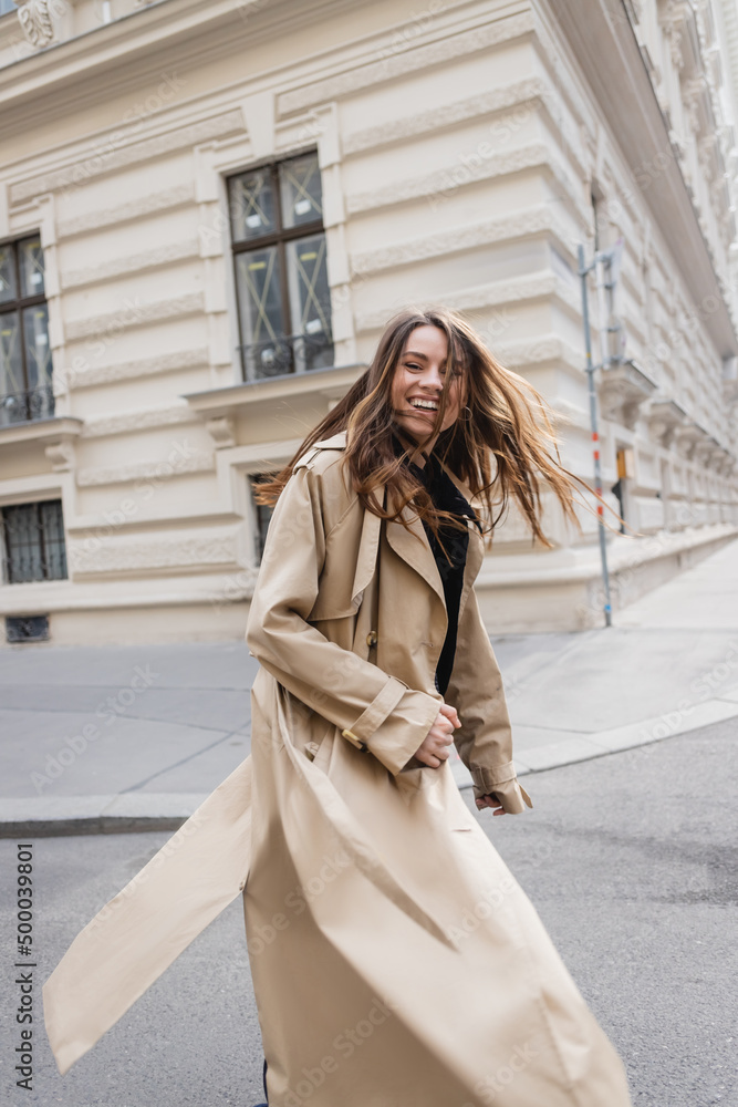 Fototapeta premium happy young woman in beige trench coat walking on european street.