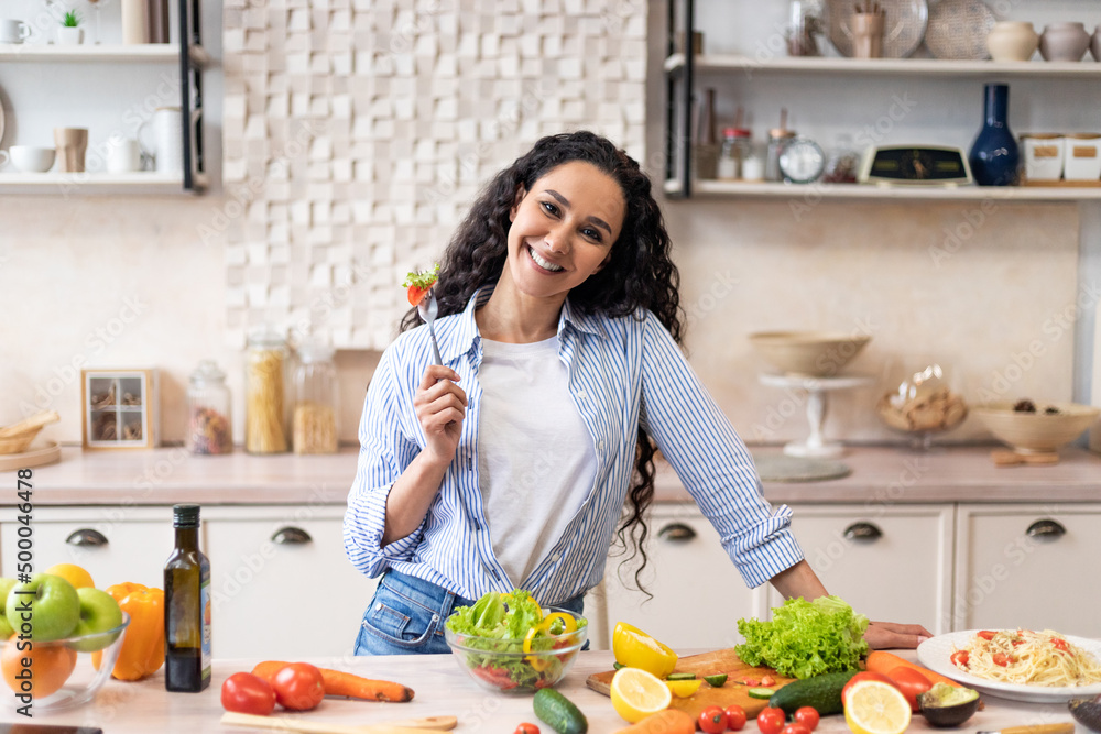Happy latin lady eating fresh vegetable salad, enjoying fresh lunch, standing in kitchen and smiling at camera