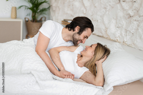 Loving young married couple relaxing, looking at each other in bed at home, feeling happy in morning