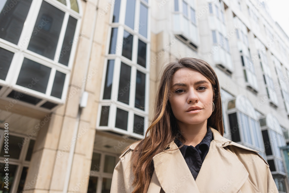 low angle view of young woman in stylish trench coat looking at camera near building.
