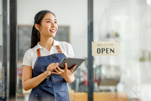 Young Asian businesswoman small business owner standing at cafeteria door entrance pen signboard, A cheerful entrepreneur young waitress in a blue apron near the glass door 