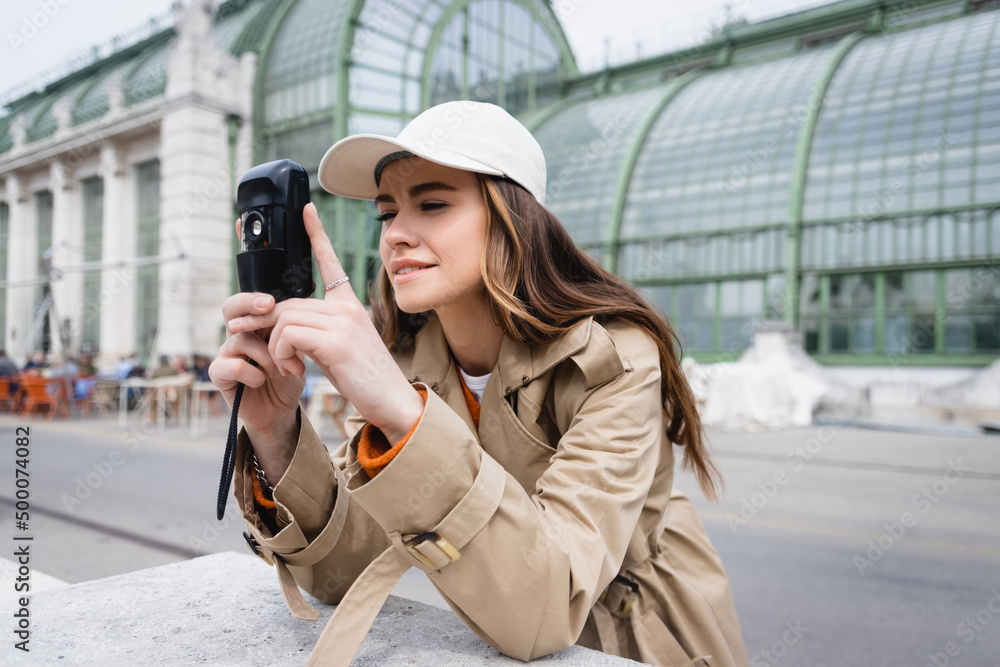 young woman in beige trench coat and baseball cap taking photo on