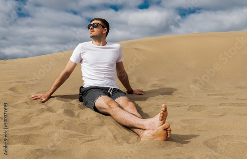 Brunette man wearing white t-shirt sitting on warm sand.