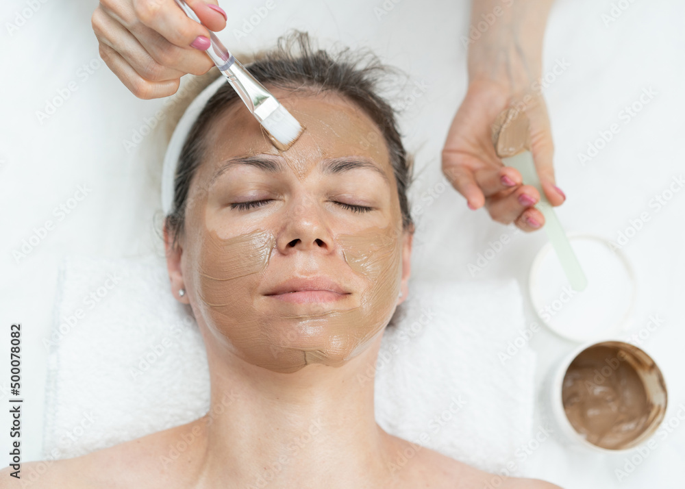 he face of a white woman undergoing cosmetic procedures. Brown clay face mask. Facial care. Applying facial mask at woman face at beauty salon A beautiful brown-haired woman enjoys applying a cosmetic
