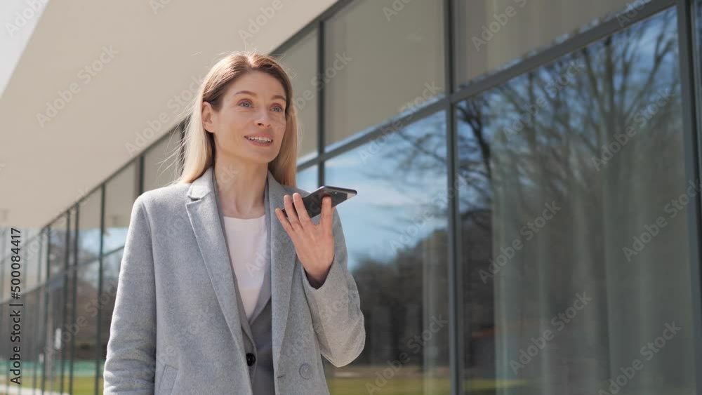 Adult happy businesswoman uses a mobile phone near the business center. Confident businesswoman holding smartphone recording voice message or activating digital assistant making business call.