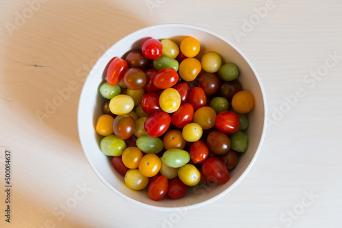 Selective focus high angle side lit flat lay view of red, yellow and green cherry tomatoes in white bowl on pale wooden background