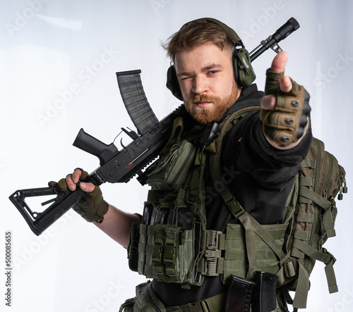 the airsoft player puts his rifle on his shoulder and aims at us with his fingers. a man in an outfit, in headphones, a bulletproof vest, with a backpack on a white background.
