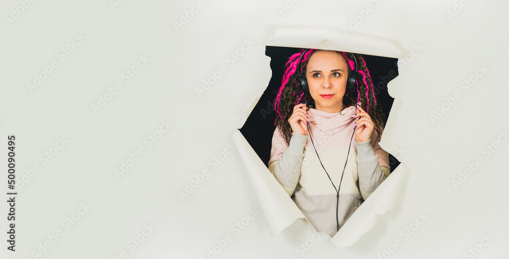 Young woman in headphones in hole of white background. Beautiful female with curly hair listening to music in studio.