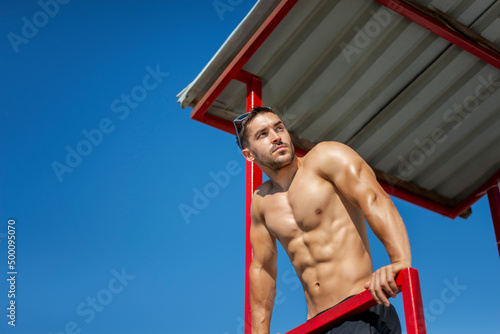 Shirtless muscular man in sunglasses on a beach.