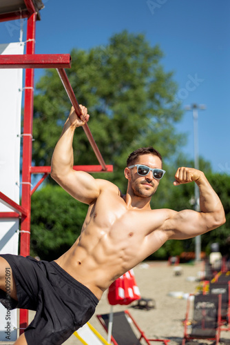 Shirtless muscular man in sunglasses on a beach.