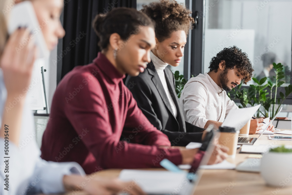 Indian businessman looking at notebook near laptop and multiethnic colleagues working in office. Indian businessman looking at notebook near laptop and multiethnic colleagues working in office.