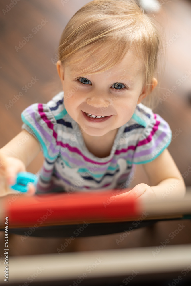 Fototapeta premium smiling happy two year old girl looking up at camera while standing next to easel magnetic board