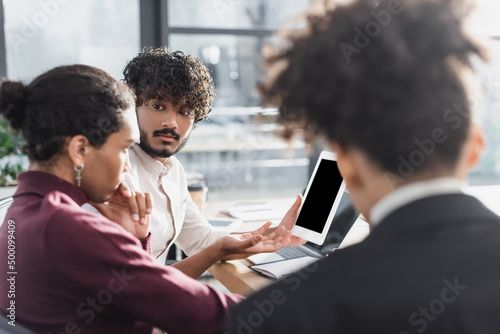 Indian businessman holding digital tablet near blurred african american colleagues in office.
