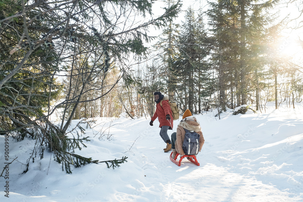 Long shot of modern young couple enjoying spending time in local forest on winter day having fun sledding