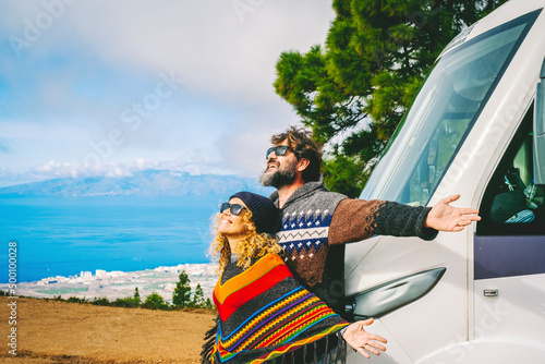 Van life and travel destination concept. Man and woman excited for new arrival in the nature opening arms against their new modern motor home camper. Free camp site with rv. People living off grid #500100028