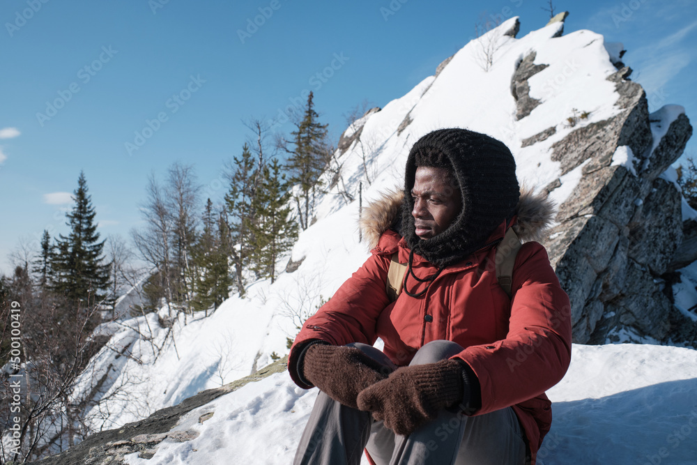 Young Black man sitting on ground covered with snow looking away on winter day somewhere in mountains