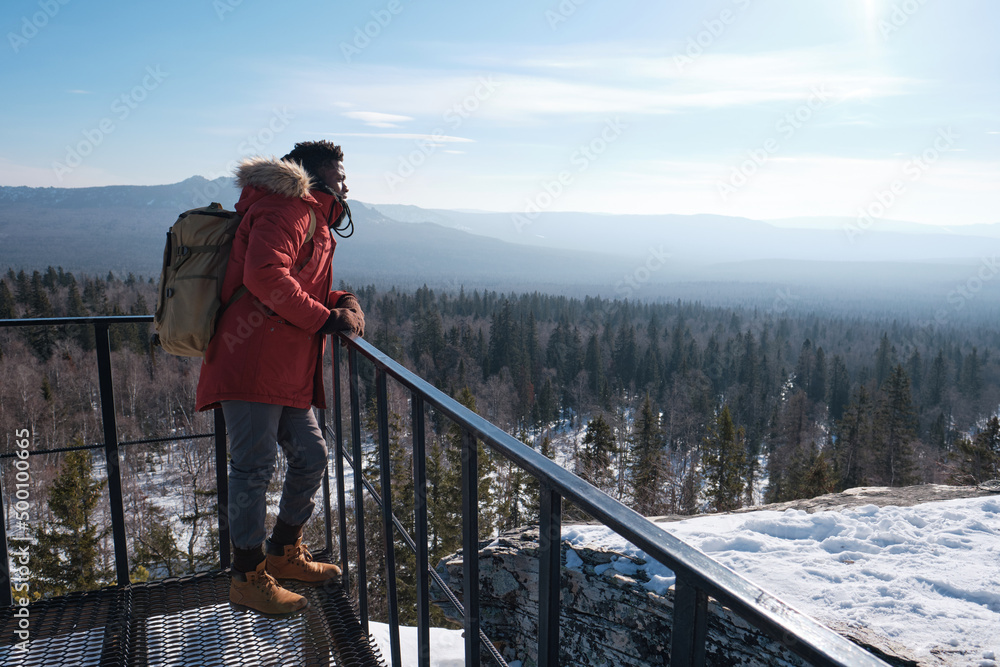 Young African American man wearing backpack standing relaxed at observation deck on mountain top on winter day Young African American man wearing backpack standing relaxed at observation deck on mountain top on winter day