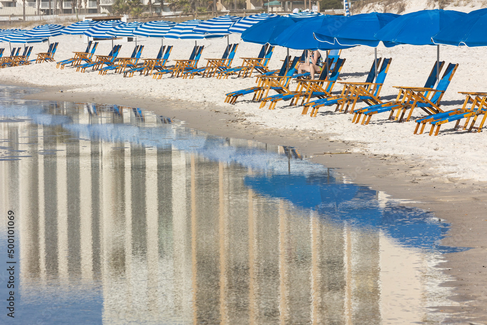 Blue and white beach chairs along the water on a white sand beach with the reflection of high-rise condos in the water.