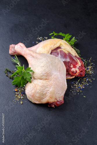 Traditional raw gourmet duck leg offered with herbs and spice as close-up on a black design board