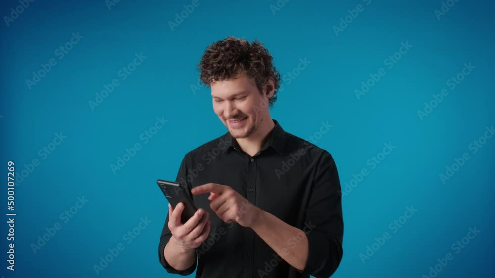 Smiling modern man chatting online message use smartphone standing isolated on blue studio Smiling modern man chatting online message use smartphone standing isolated on blue studio