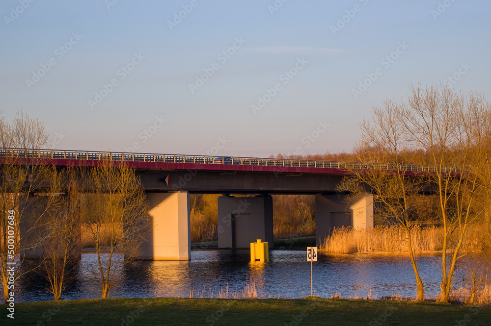 Pięknie oświetlony most nad rzeką w otoczeniu żółtych traw - obrazy, fototapety, plakaty 