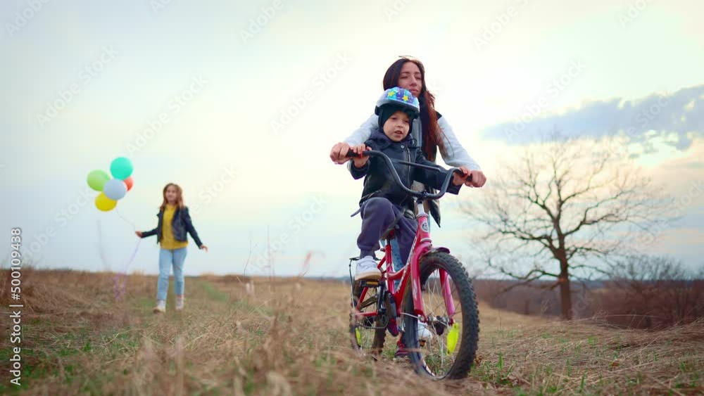 Childhood bike concept. Mother teaching son to ride bicycle. Young mom teaching son to ride bike first time on countryside rural road at sunset. Happy kids on bikes. The best moments together. Childhood bike concept. Mother teaching son to ride bicycle. Young mom teaching son to ride bike first time on countryside rural road at sunset. Happy kids on bikes. The best moments together.