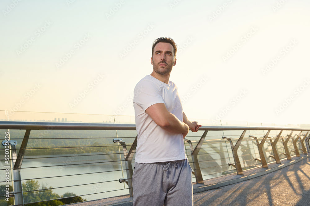 Middle aged European man in sportswear exercising outdoor on the city bridge at dawn on a warm sunny summer day. Sport, active and healthy lifestyle concept. Keep you body fit Middle aged European man in sportswear exercising outdoor on the city bridge at dawn on a warm sunny summer day. Sport, active and healthy lifestyle concept. Keep you body fit
