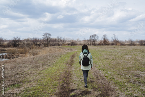 Fototapeta Naklejka Na Ścianę i Meble -  A girl with a backpack walks along the road in a green field, a spring walk through the park in nature, a hike in the forest in the spring.
