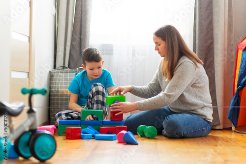 Mother with her son playing with colored cubes sitting on the floor at home. Family spends time together at home in children's room