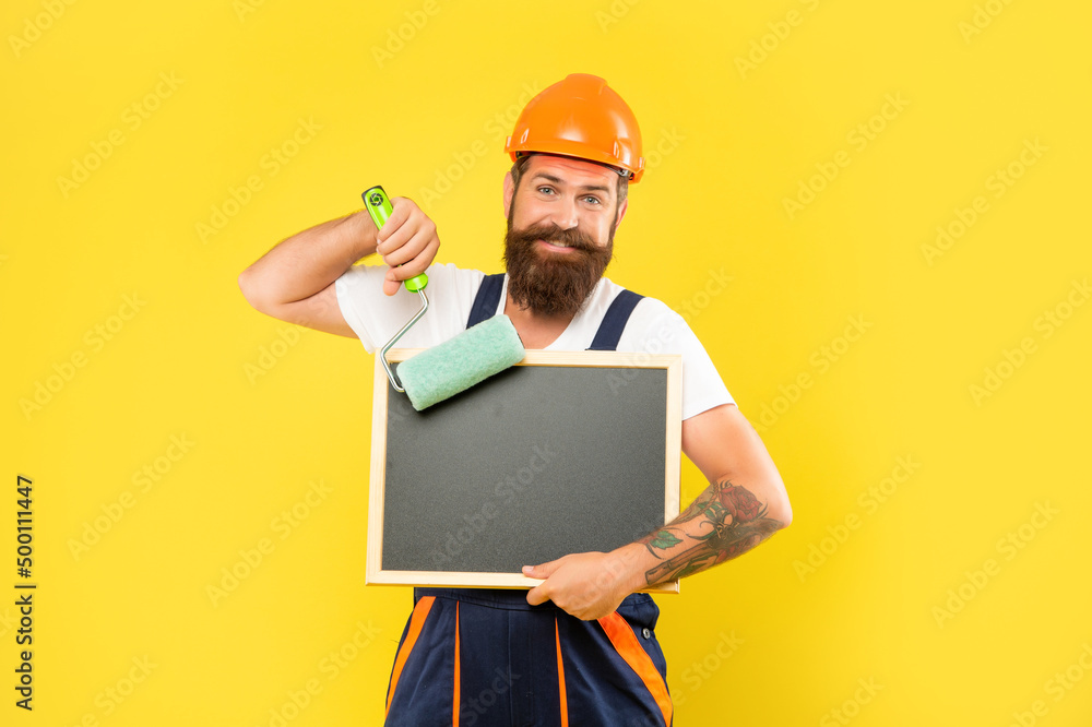 positive man decorator in helmet and work clothes hold paint roller and blackboard with copy space positive man decorator in helmet and work clothes hold paint roller and blackboard with copy space