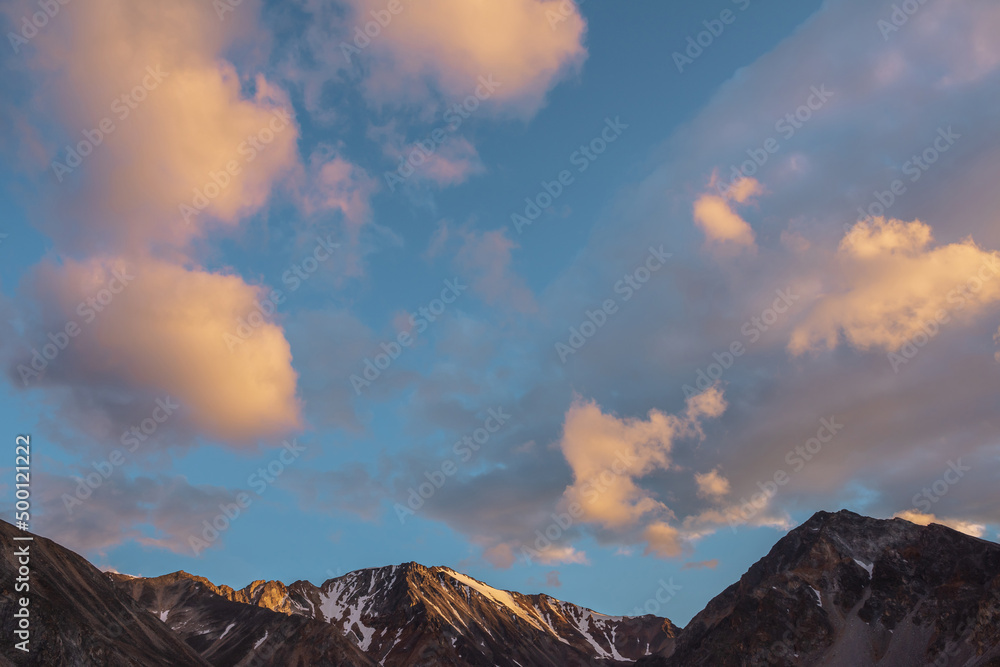 Scenic landscape with high snow mountain with sharp rocky pinnacle in golden sunlight under clouds of sunset color at changeable weather. Colorful view to large mountain top under orange clouds in sky