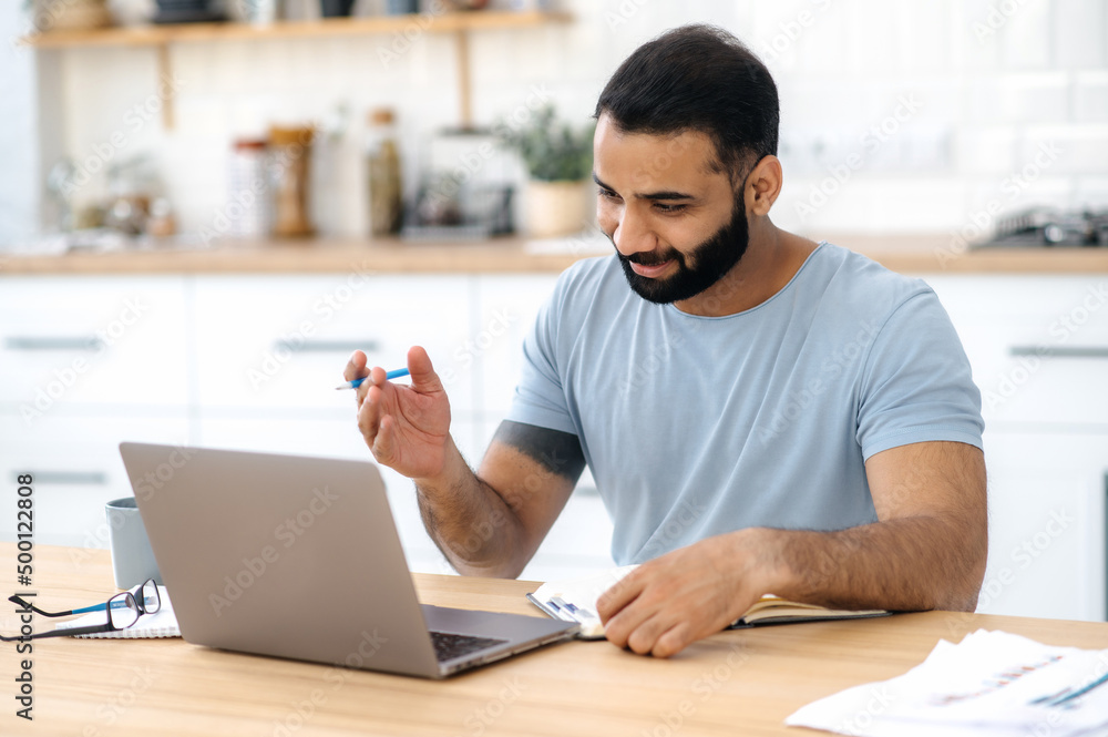 Positive modern Indian man, freelancer, designer or student, sitting at a kitchen at home, studying or working online uses laptop, smiling friendly, takes notes. E-learning, video conference concept