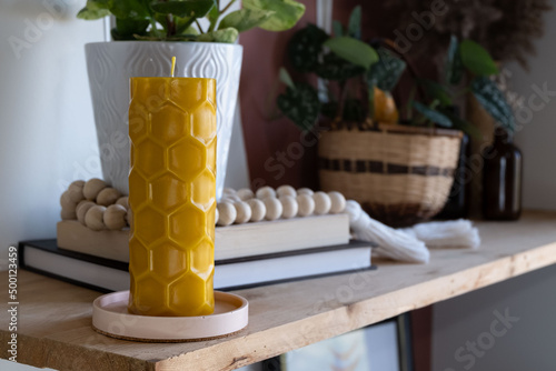 A beeswax pillar candle with a hex design is displayed on a shelf with various decor in the background.