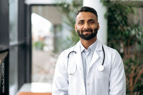 Portrait of a handsome Indian general therapist doctor, in medical uniform and stethoscope, standing in the hospital, looking at the camera, smiling friendly