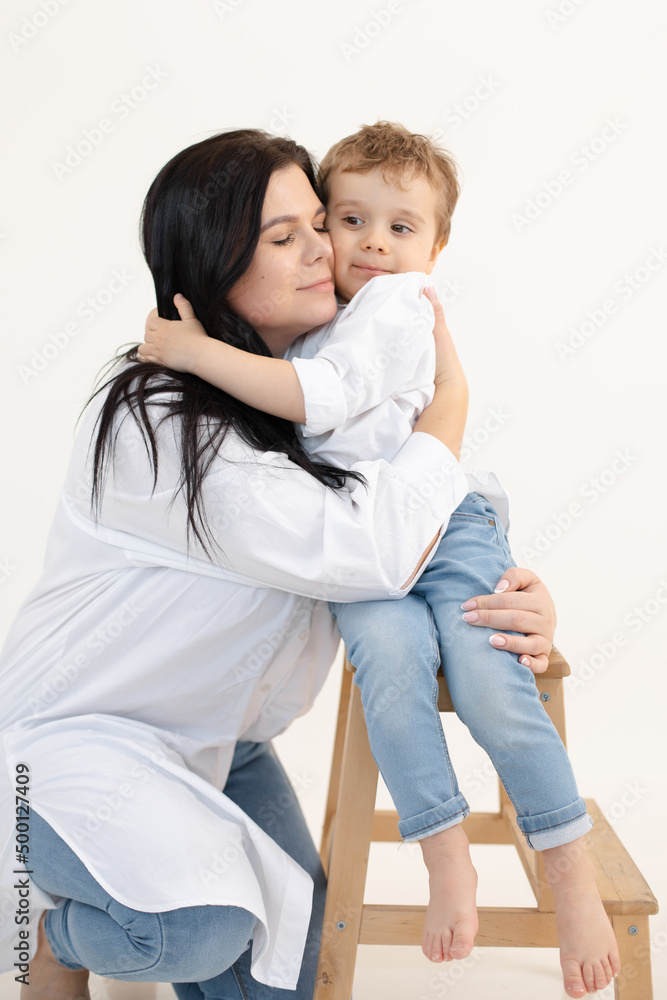 Vertical photo of brunette glad mom and son embracing, boy sitting on wooden chair in studio. Spending time together