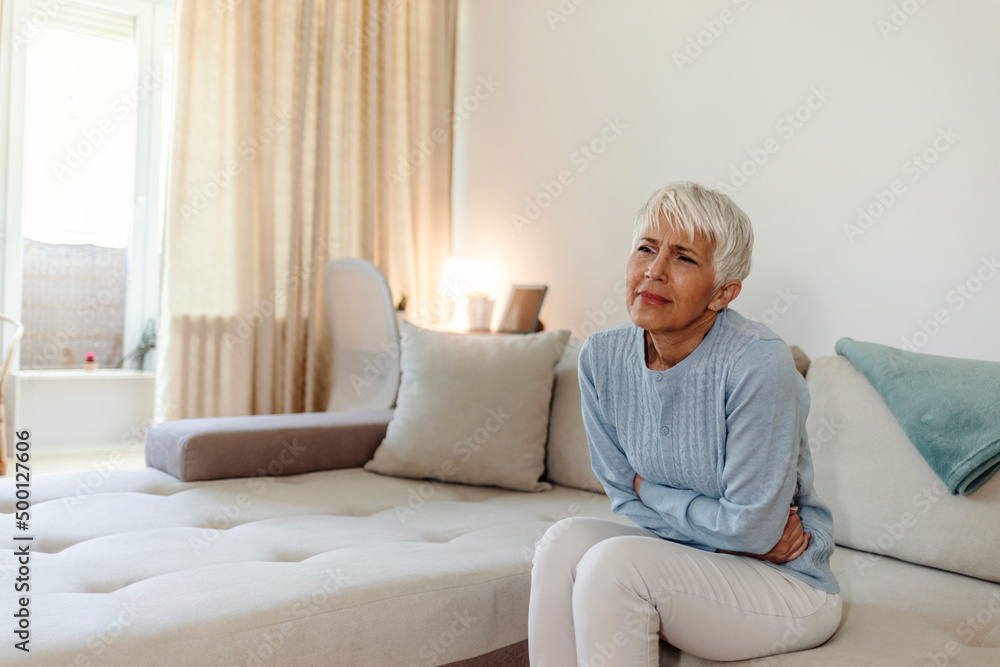 Mature woman suffering from stomach ache sitting on a bed in the bedroom. Photo of mature adult woman sitting on her bed has a stomach ache in the bedroom. Senior woman suffering from stomach pain Mature woman suffering from stomach ache sitting on a bed in the bedroom. Photo of mature adult woman sitting on her bed has a stomach ache in the bedroom. Senior woman suffering from stomach pain