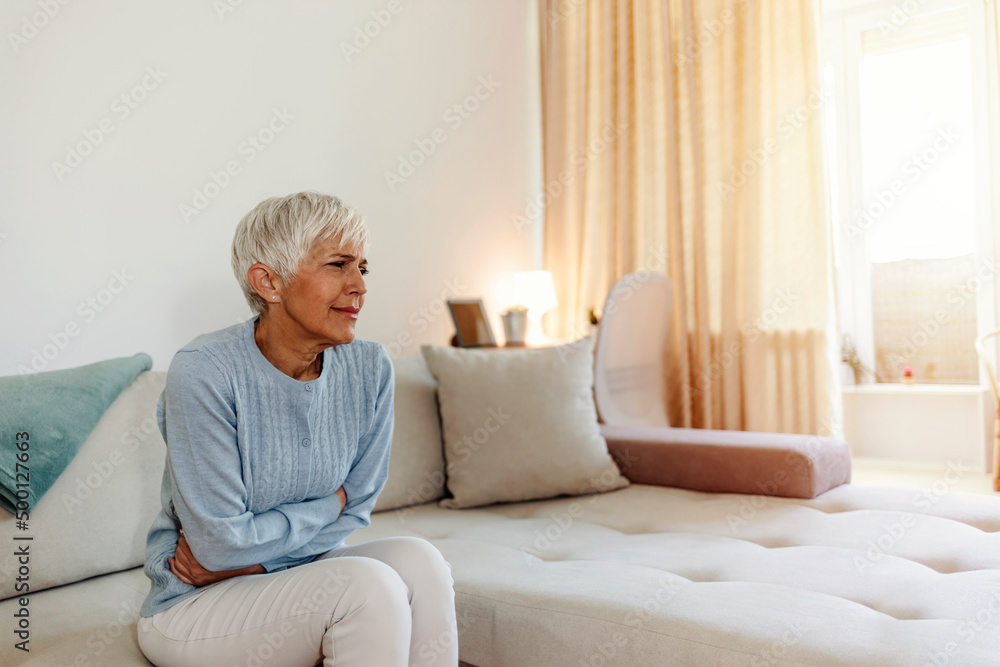 Mature woman suffering from stomach ache sitting on a bed in the bedroom. Photo of mature adult woman sitting on her bed has a stomach ache in the bedroom. Senior woman suffering from stomach pain Mature woman suffering from stomach ache sitting on a bed in the bedroom. Photo of mature adult woman sitting on her bed has a stomach ache in the bedroom. Senior woman suffering from stomach pain
