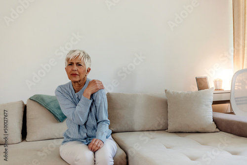 Stressed mature woman massaging her neck. Mature woman suffering from neck pain at home. Portrait of elderly woman having a neck pain in the living room. Stress and depression. © Jelena Stanojkovic