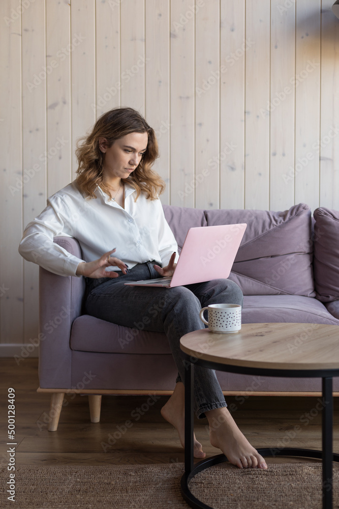 Portrait of woman with bare foot working on laptop sitting on soft violet sofa in casual clothes near table with cup of coffee. Living room in elegant design with aesthetic decor, free copy space Portrait of woman with bare foot working on laptop sitting on soft violet sofa in casual clothes near table with cup of coffee. Living room in elegant design with aesthetic decor, free copy space