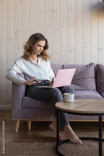 Free Download Portrait of woman with bare foot working on laptop sitting on soft violet sofa in casual clothes near table with cup of coffee. Living room in elegant design with aesthetic decor, free copy space - GFXLoad