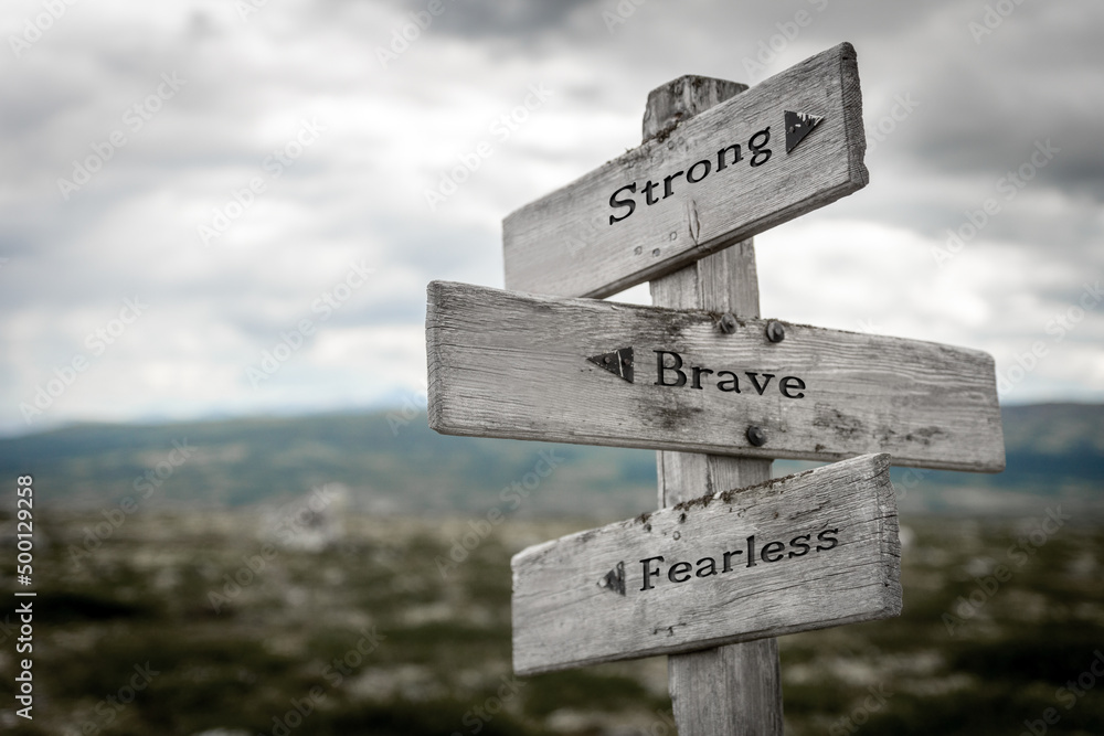 strong brave fearless text quote written in wooden signpost outdoors in nature. Moody theme feeling.