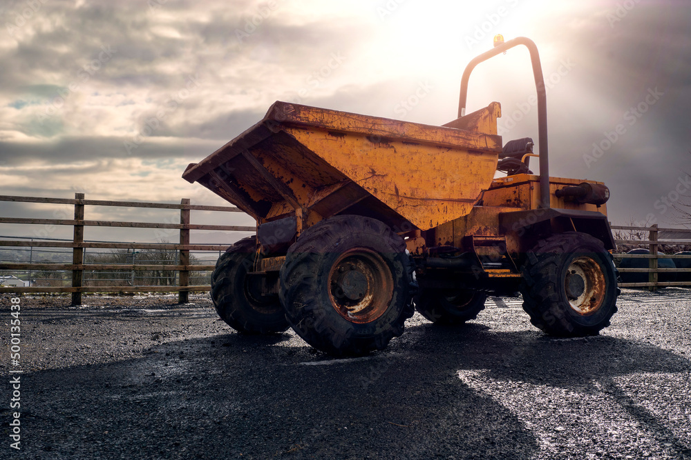 Obraz premium Old rusty dumper track parked on a parking lot in a country side. The vehicle is yellow color has many scratches. Warm sunny day. Dramatic sky. Construction and repair equipment for rent concept.