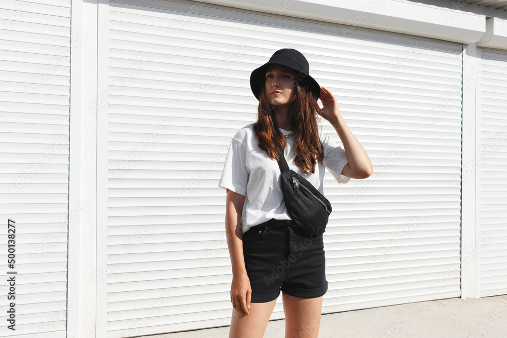 Woman, wearing white t-shirt, black shorts, fanny pack or waist pack and bucket  hat, standing outdoor near white wall. Details of stylish trendy basic  minimalistic casual outfit. Street fashion. Photos