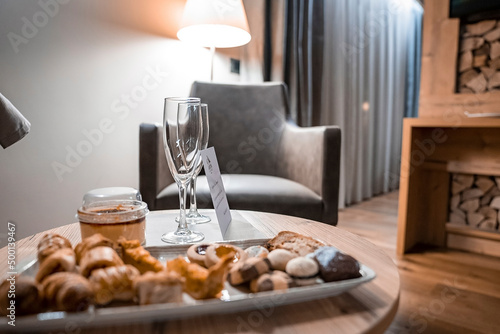 Sweet baked food served in dish by champagne glasses and greeting card on table. Armchair and illuminated lamp in background. Interior of rustic room in luxurious resort. photo