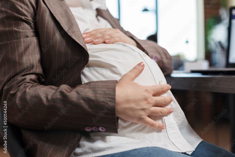 Obraz premium Close up of pregnant woman touching baby bump while in company office workplace. Future young mother sitting in marketing company workplace while holding baby belly bump.
