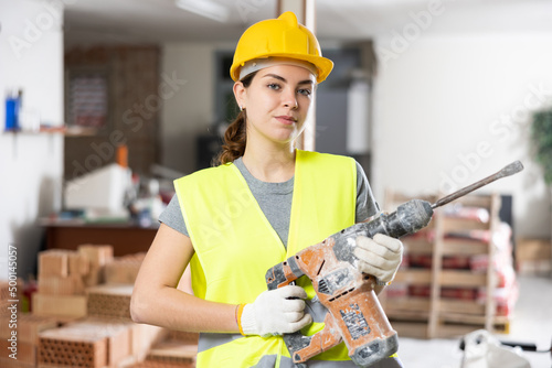 Young woman builder standing with pneumatic hammer drill on indoor building construction site and smiling photo
