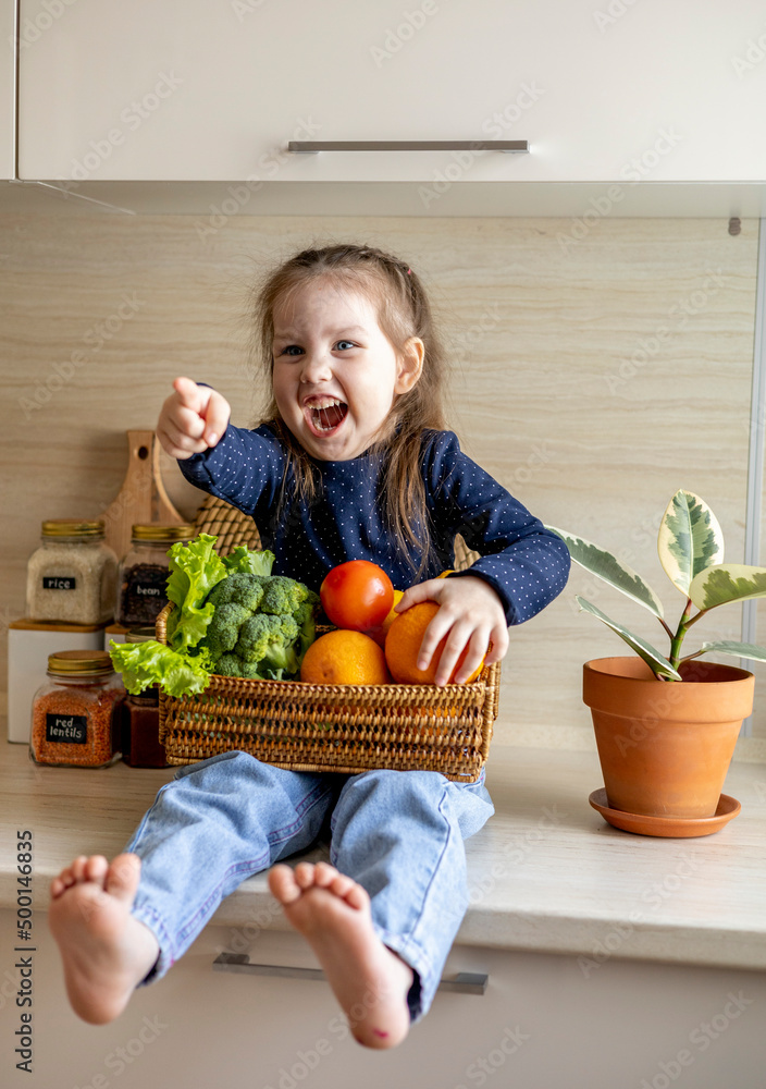 Fototapeta premium Baby sits on the kitchen table and holds a wicker basket of fruit in her hands.